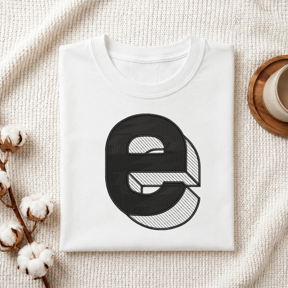 A neatly folded white crewneck t-shirt featuring a stitched 3D block lowercase letter "e" design, styled with cotton branches and a wooden coaster.