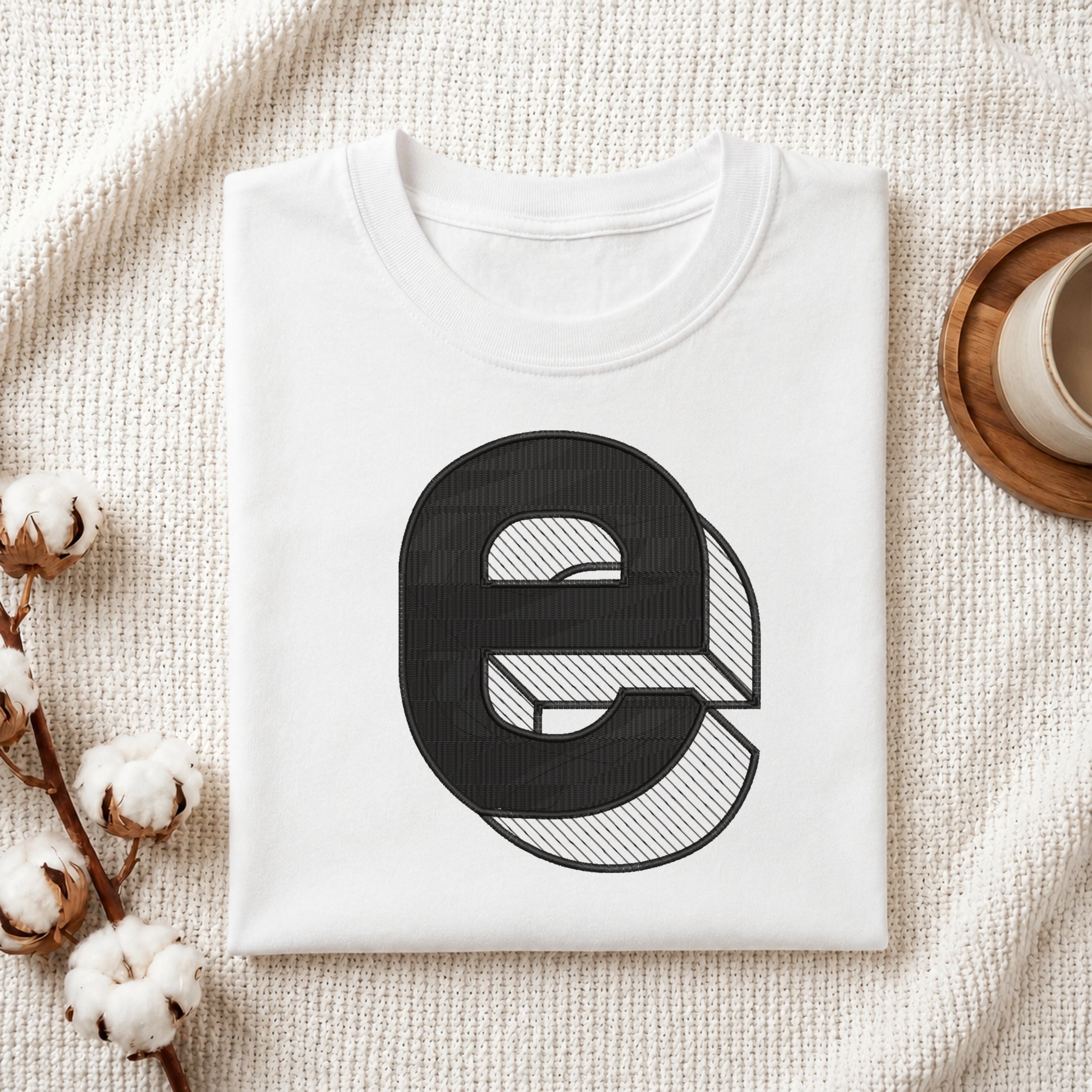 A neatly folded white crewneck t-shirt featuring a stitched 3D block lowercase letter "e" design, styled with cotton branches and a wooden coaster.