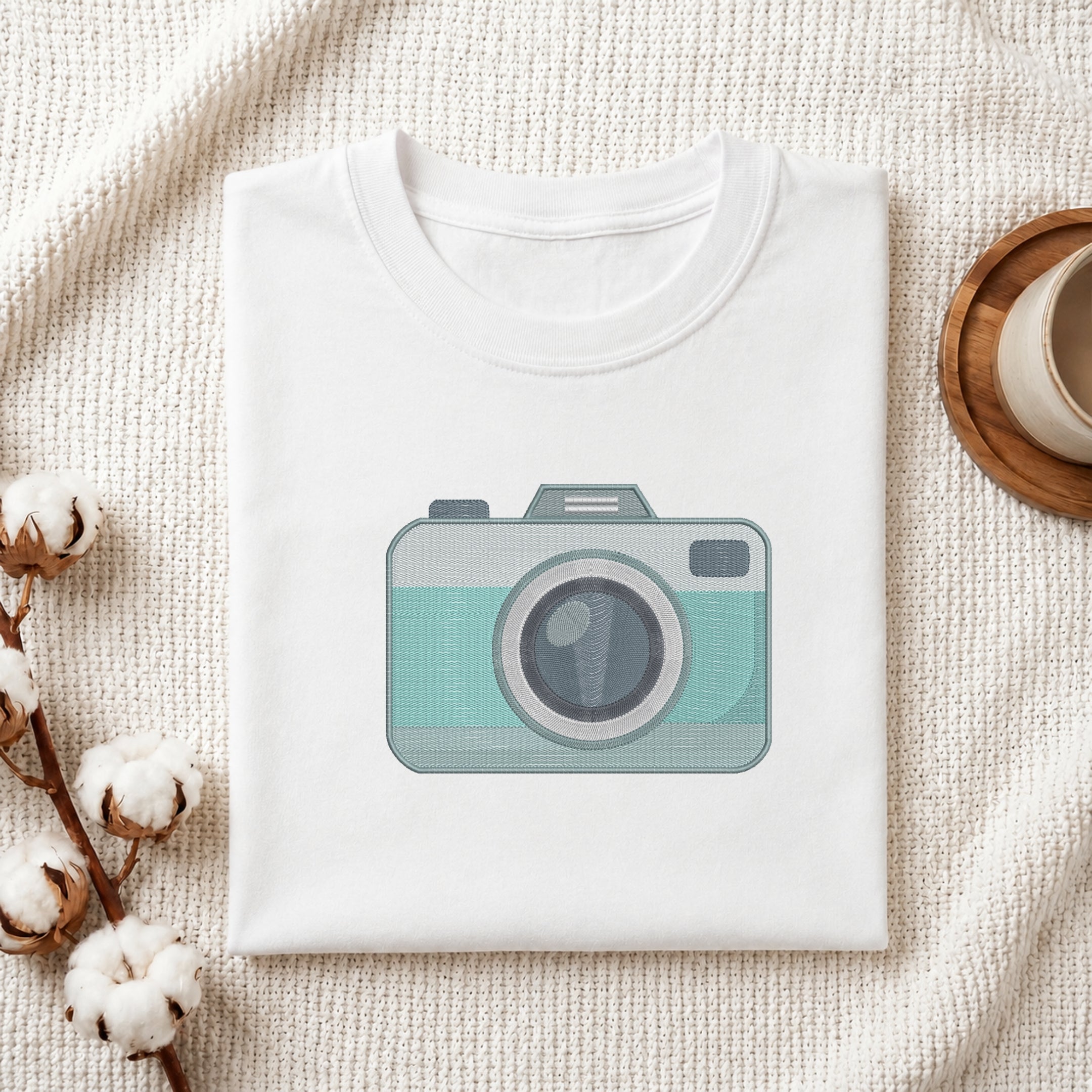 A folded white crewneck t-shirt with a graphic of a retro camera in mint green and gray, styled on a textured cream knit blanket with cotton bolls and a wooden cup.