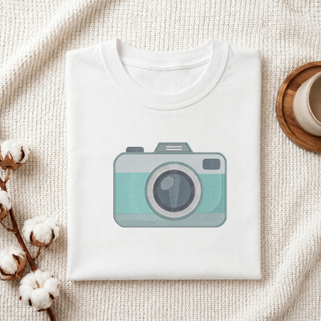 A folded white crewneck t-shirt with a graphic of a retro camera in mint green and gray, styled on a textured cream knit blanket with cotton bolls and a wooden cup.