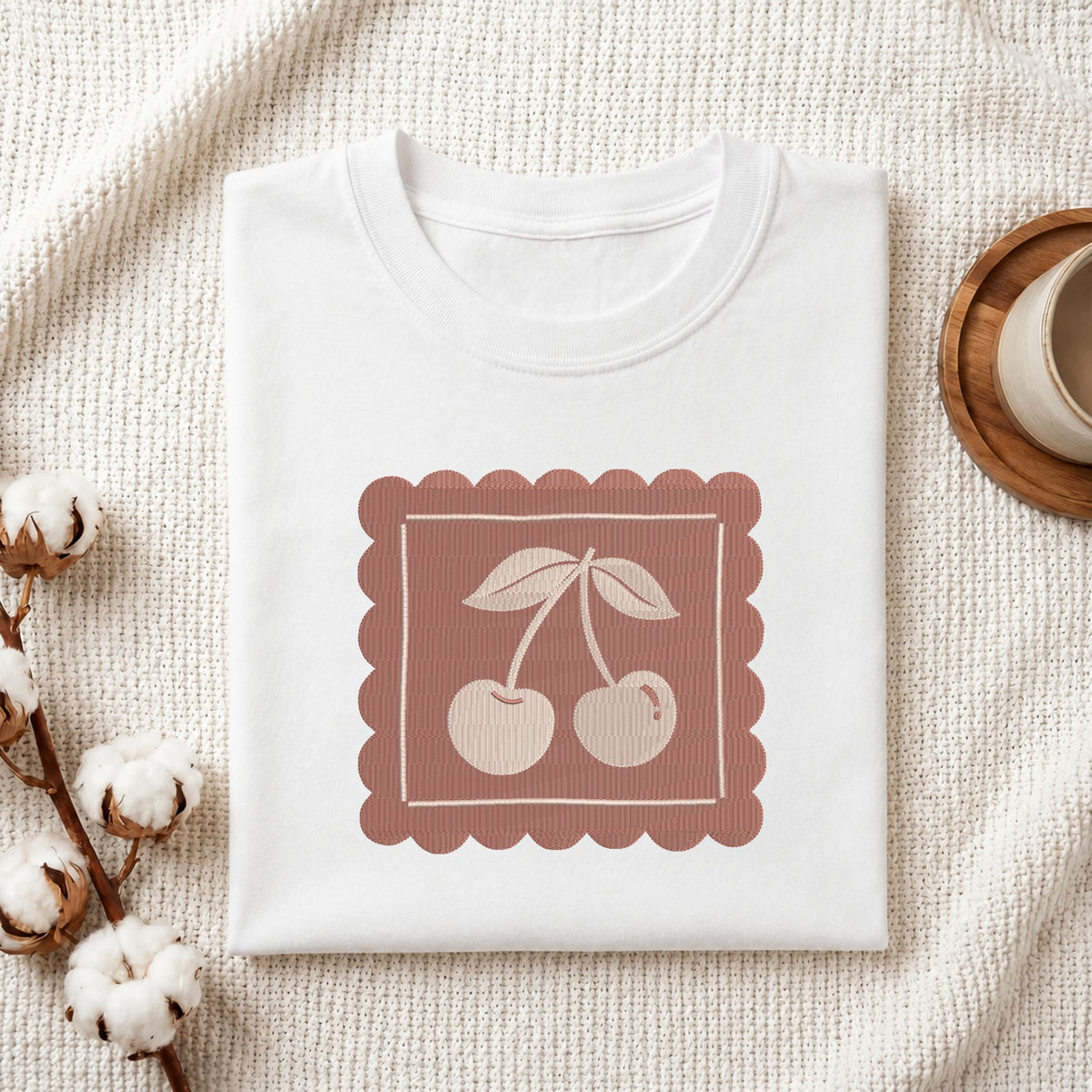 Folded white cotton t-shirt mockup featuring a vintage cherry machine embroidery design, styled with cotton stems and a wooden cup