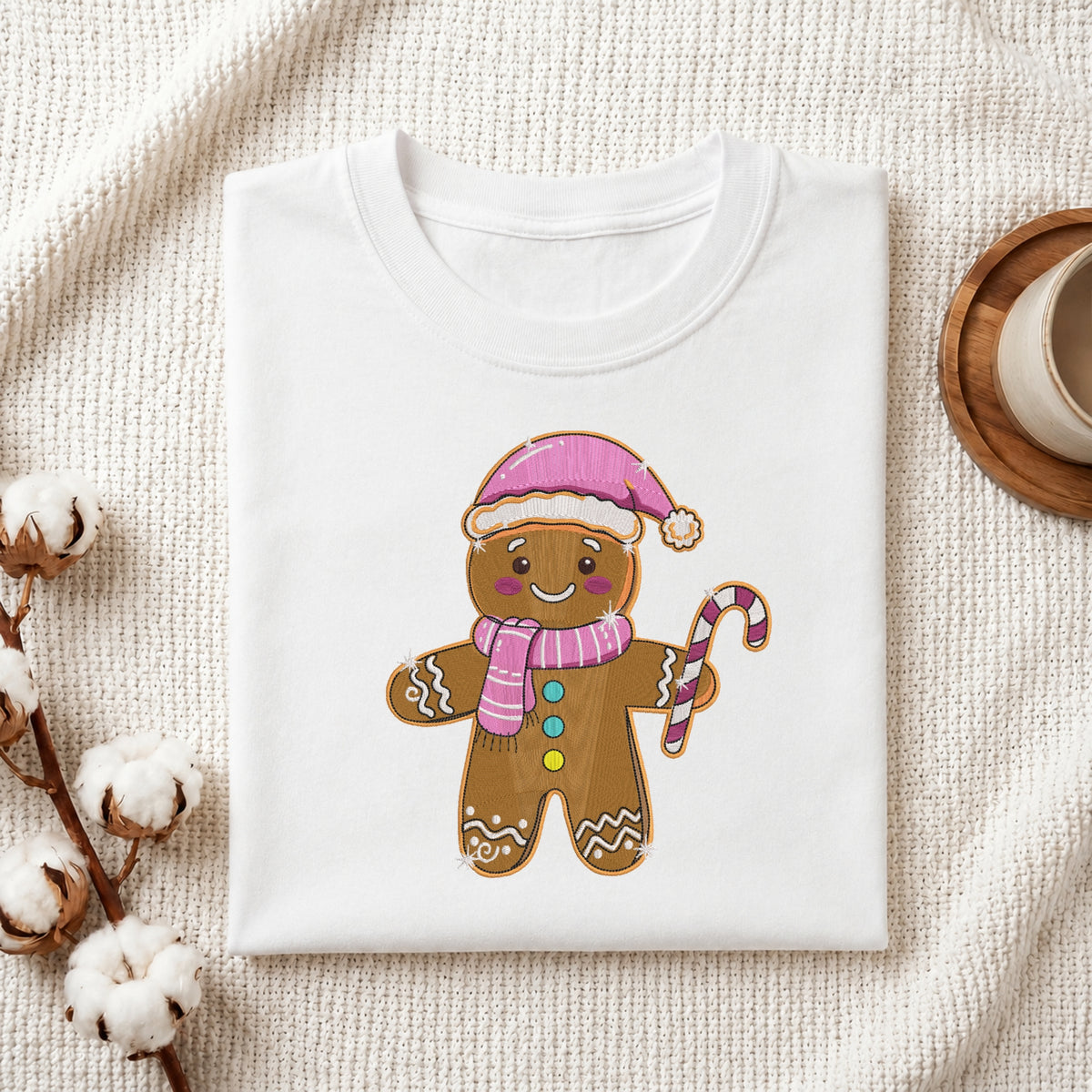 A folded white crewneck t-shirt featuring a cute gingerbread man graphic with a pink Santa hat, pink striped scarf, and candy cane, styled on a textured cream fabric with cotton stems and a wooden cup.
