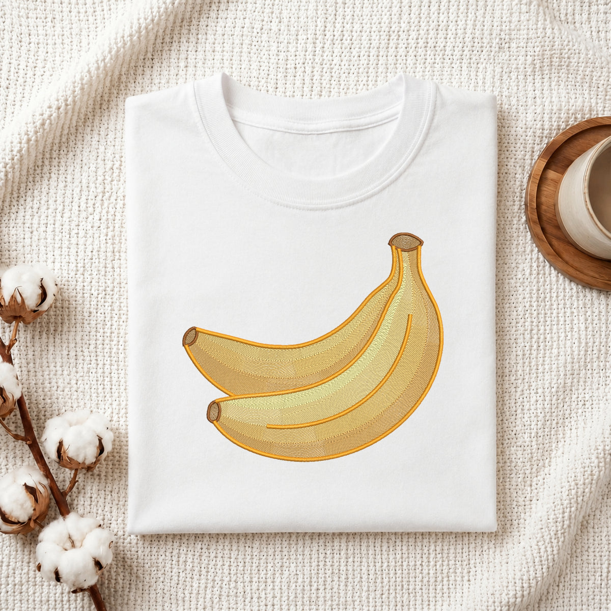 A folded white crewneck T-shirt with a graphic of two yellow bananas, styled on a textured cream fabric with cotton stems and a wooden cup in the background.