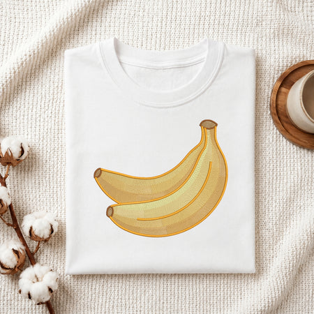 A folded white crewneck T-shirt with a graphic of two yellow bananas, styled on a textured cream fabric with cotton stems and a wooden cup in the background.