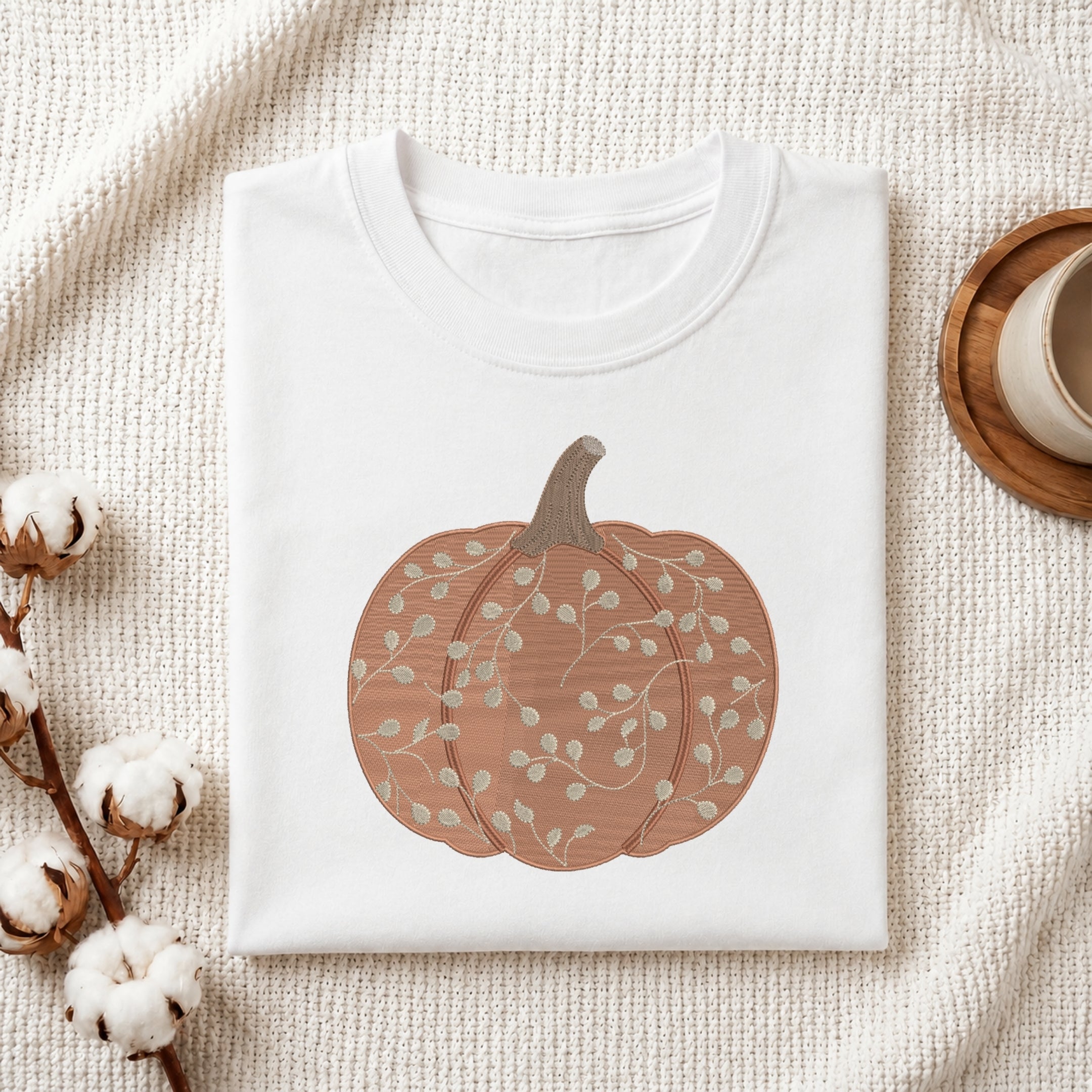 A folded white crewneck t-shirt with a graphic of a brown and beige autumn pumpkin, styled on a textured cream fabric with cotton bolls and a wooden cup in the background.