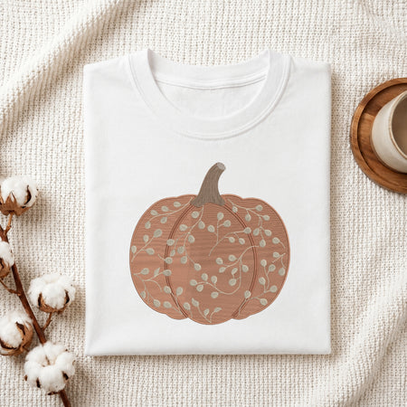A folded white crewneck t-shirt with a graphic of a brown and beige autumn pumpkin, styled on a textured cream fabric with cotton bolls and a wooden cup in the background.