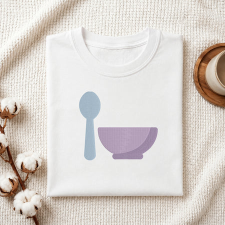 A folded white crewneck t-shirt with a minimalist graphic of a light blue spoon and a lavender purple bowl, styled on a textured cream knit blanket with cotton bolls and a wooden cup in the background.