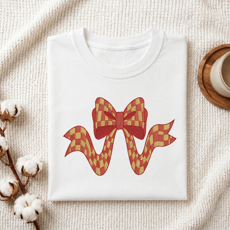 A folded white crewneck t-shirt with a red and yellow checkered bow graphic on the front, styled on a textured cream fabric background with cotton stems and a wooden cup.