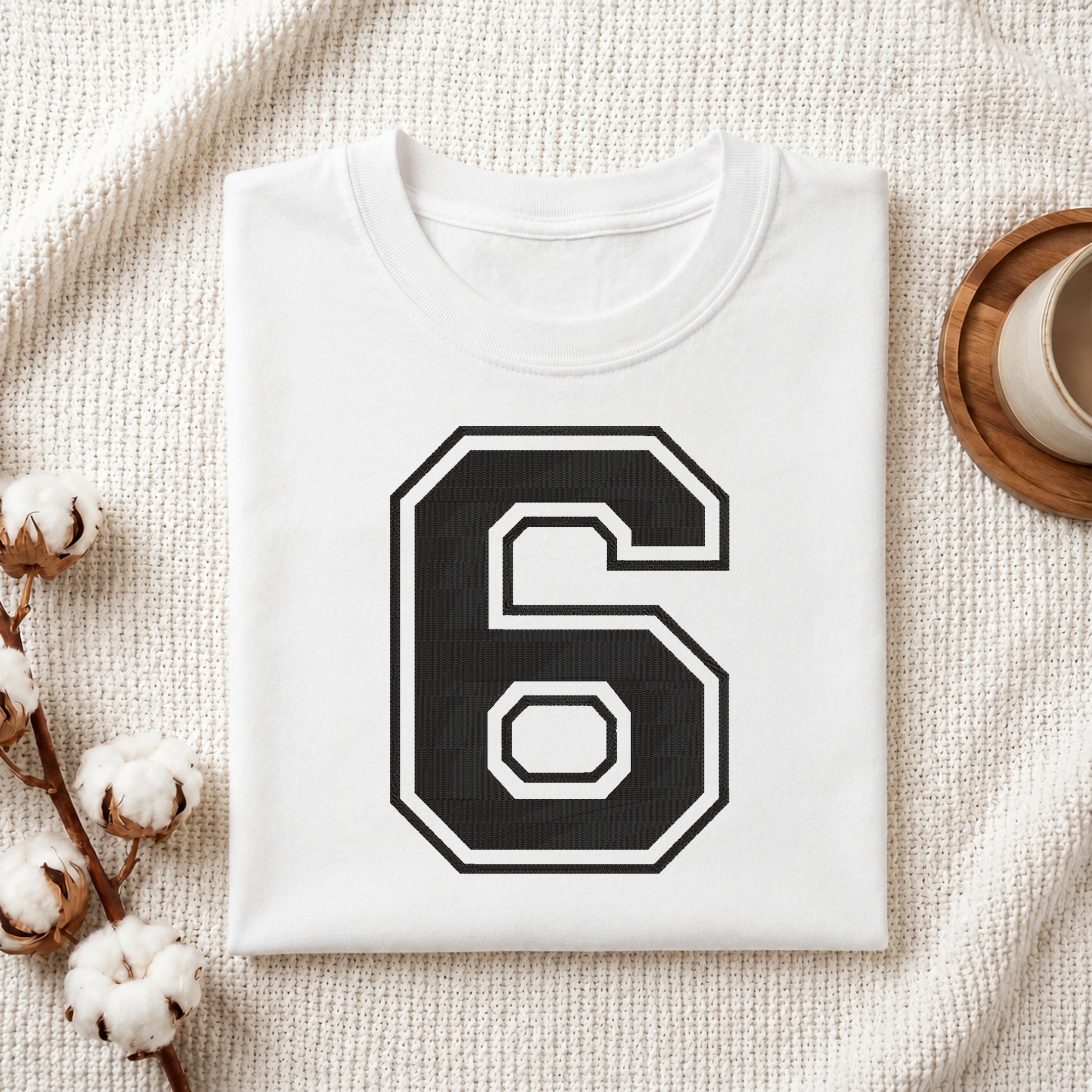 A folded white t-shirt with a large black and white number 6 printed on the front, styled on a textured cream fabric with cotton bolls and a wooden cup in the background.