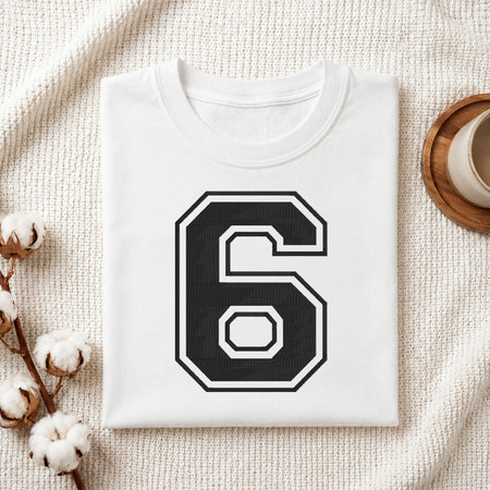 A folded white t-shirt with a large black and white number 6 printed on the front, styled on a textured cream fabric with cotton bolls and a wooden cup in the background.
