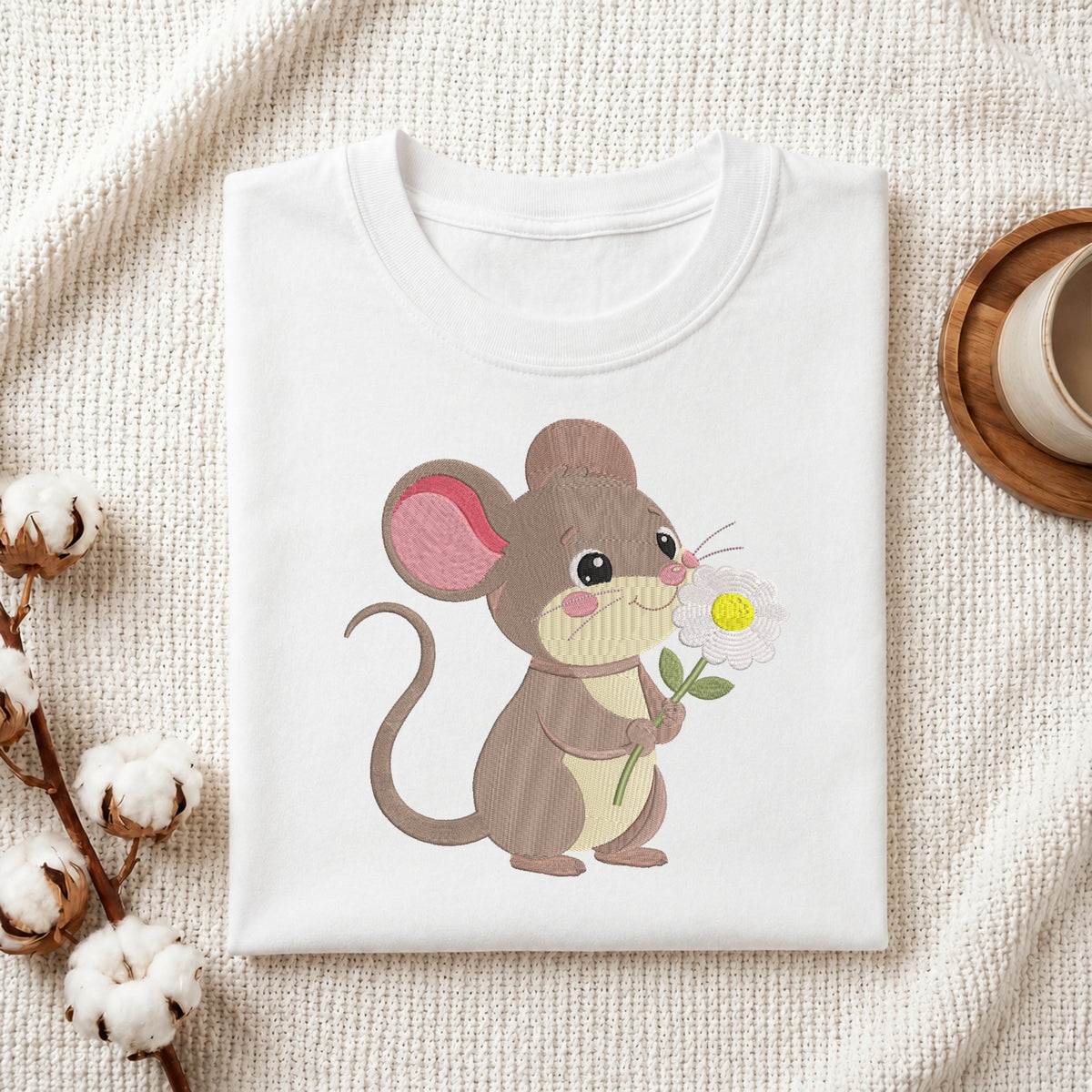 A folded white T-shirt with a cute embroidered-style mouse holding a daisy, displayed on a textured cream fabric with cotton stems and a small cup in the background.