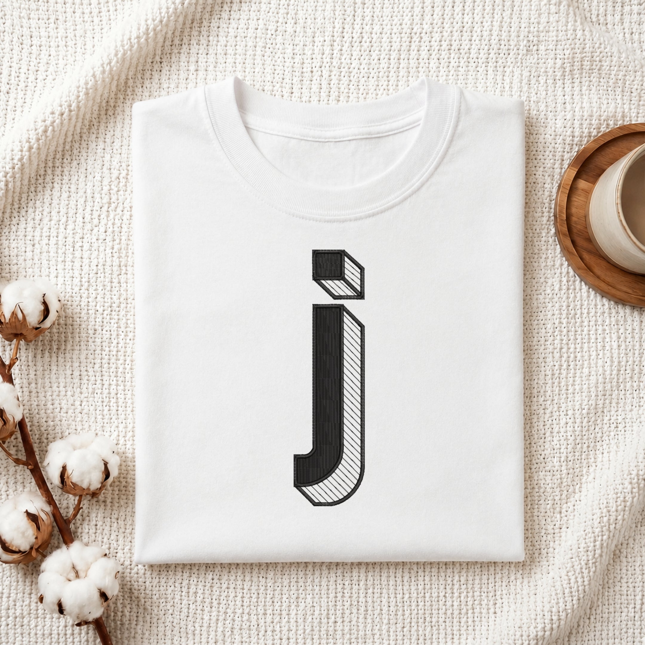 A folded white t-shirt with a black stylized lowercase letter 'j' printed on the front, displayed in a flat-lay arrangement on a textured cream fabric with cotton bolls and a wooden cup.