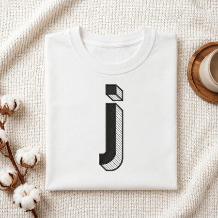A folded white t-shirt with a black stylized lowercase letter 'j' printed on the front, displayed in a flat-lay arrangement on a textured cream fabric with cotton bolls and a wooden cup.