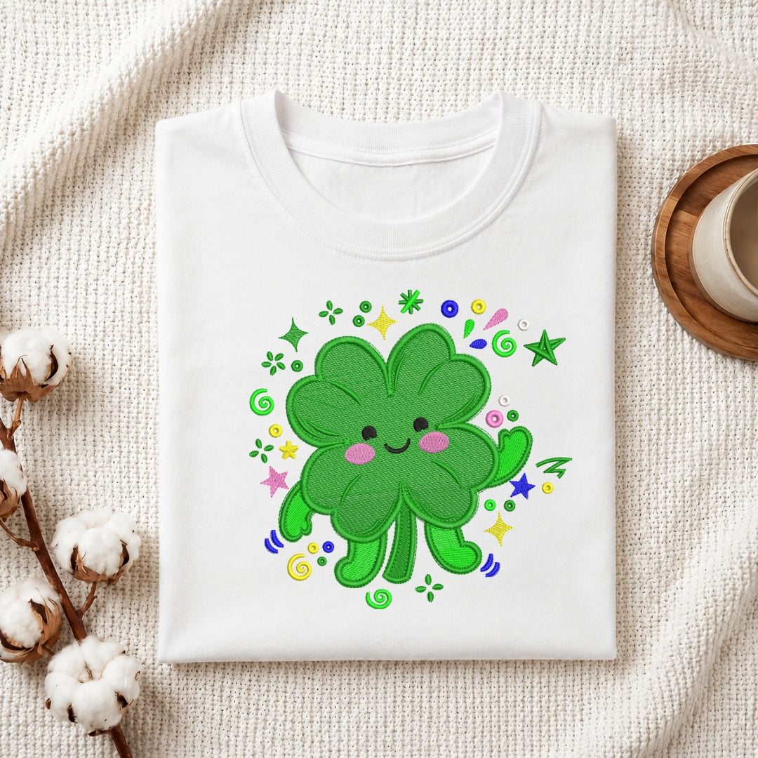 A folded white T-shirt displayed on a textured cream fabric background, decorated with cotton bolls on the left and a wooden cup on the right. The shirt features a colorful embroidered design of a smiling green shamrock with pink cheeks and small multicolored stars and swirls.