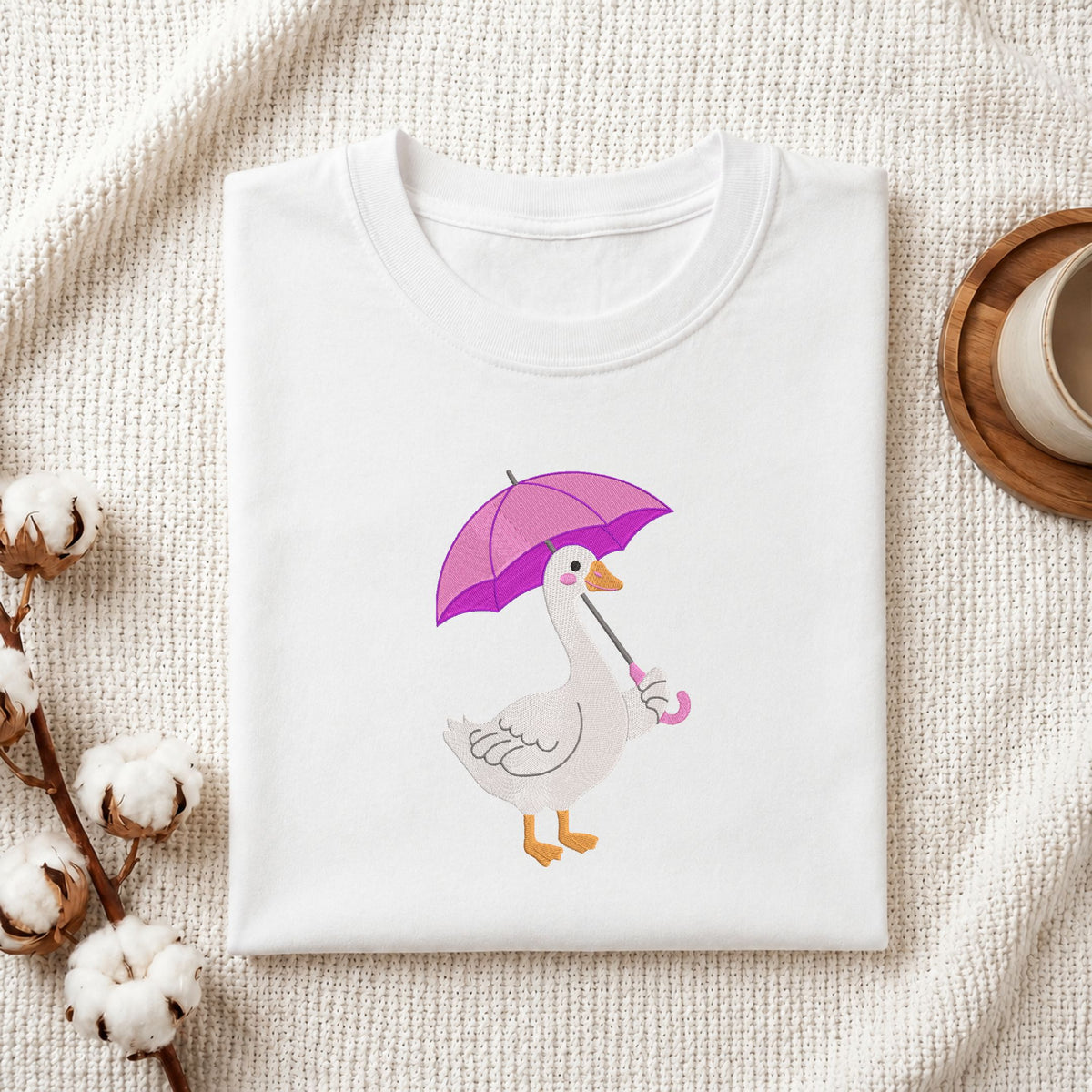 Folded white cotton t-shirt mockup featuring a cute duck with a purple umbrella machine embroidery design, styled with cotton stems and a wooden cup.   