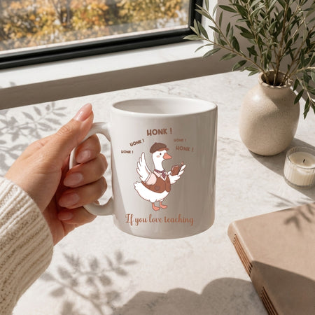 Hand holding a white ceramic coffee mug printed with the funny "HONK! If you love teaching" duck illustration.