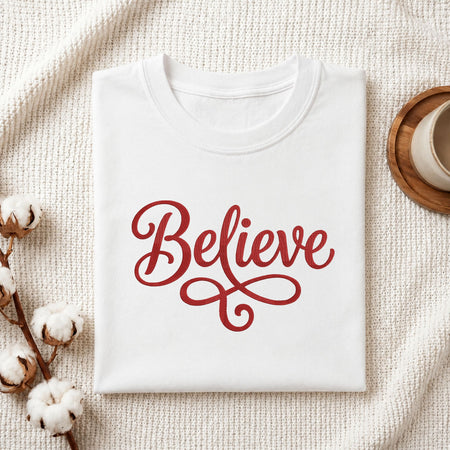 Folded white cotton t-shirt mockup featuring a "Believe" cursive embroidery design, styled with cotton stems and a wooden cup.