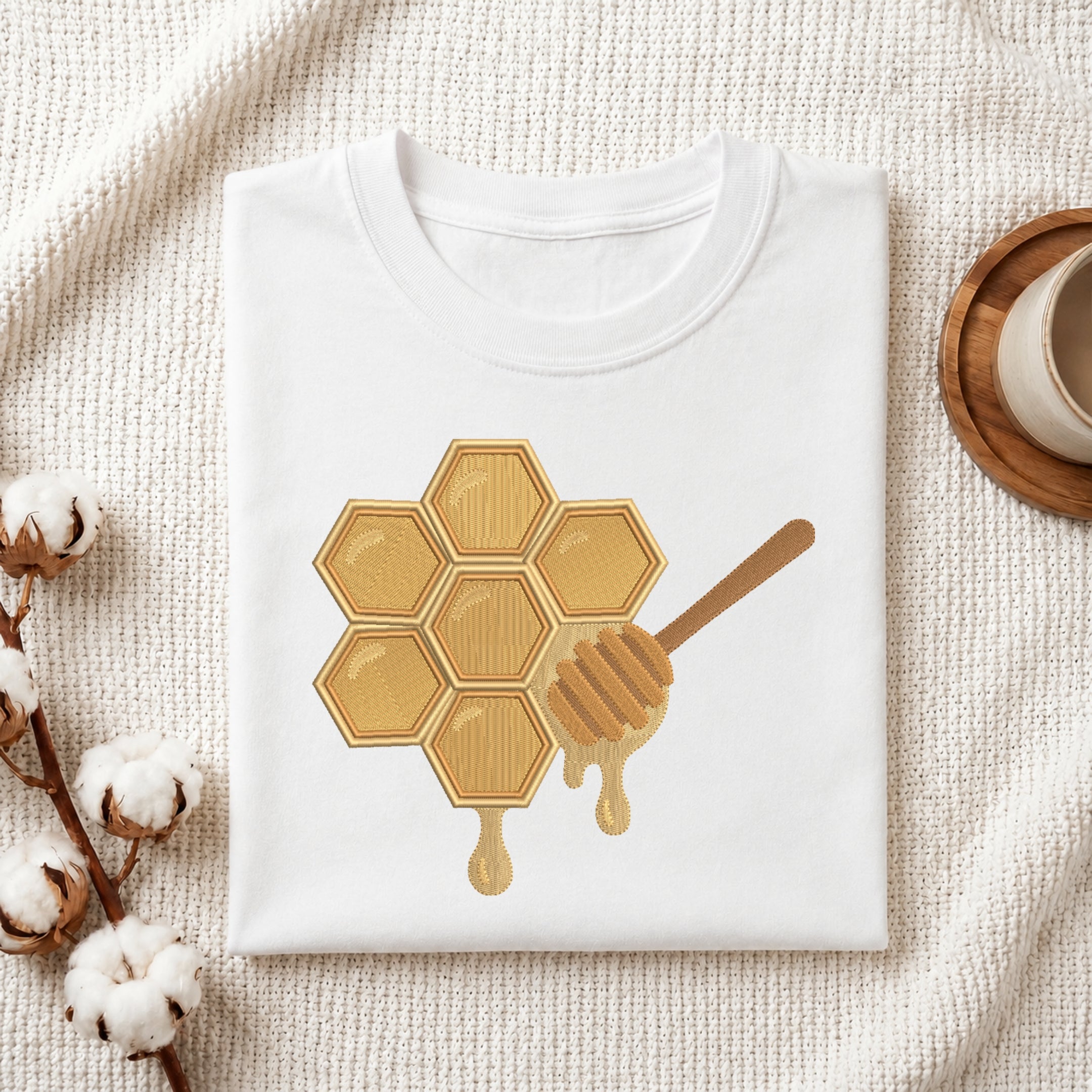 A folded white T-shirt with a honeycomb and honey dipper graphic in warm gold and brown tones, styled on a textured cream fabric with cotton bolls and a small cup at the side.