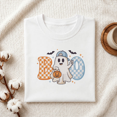 A folded white t-shirt with a Halloween graphic featuring a cartoon ghost, a jack-o'-lantern, and the word 'BOO' in orange and blue checkerboard patterns, styled on a textured cream fabric with cotton stems and a wooden cup.