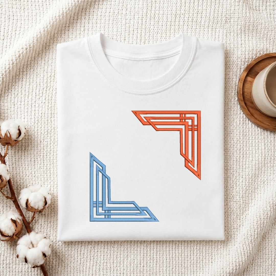 A folded white crewneck t-shirt with two geometric corner designs (one orange, one blue) on the front, styled on a textured cream blanket with cotton bolls and a coffee cup.