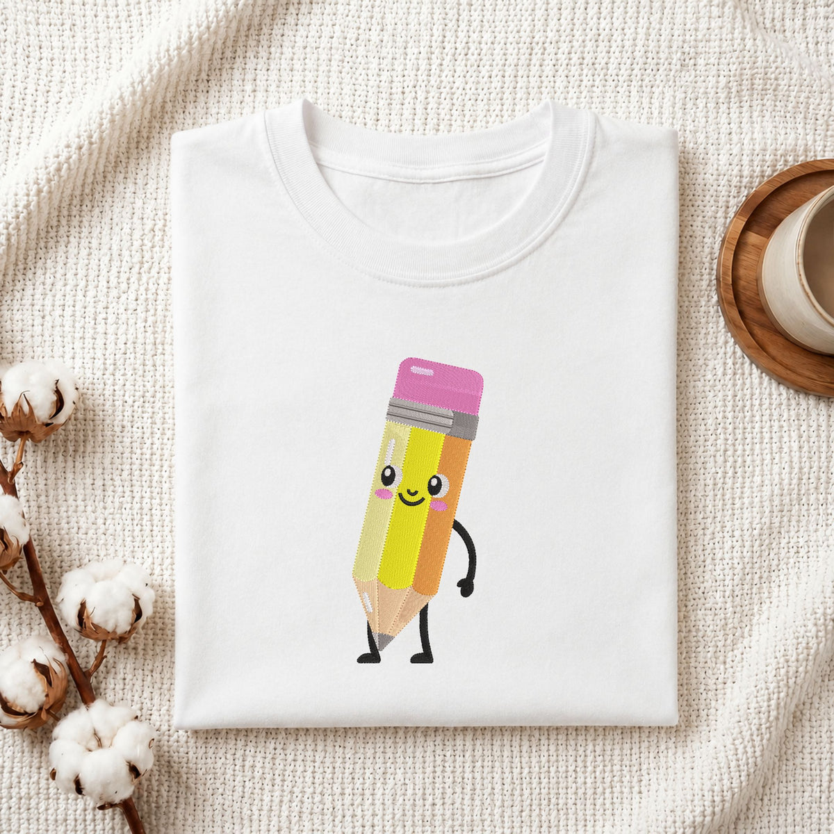 Folded white cotton t-shirt mockup featuring a cute cartoon pencil character machine embroidery design, styled with cotton stems and a wooden cup.