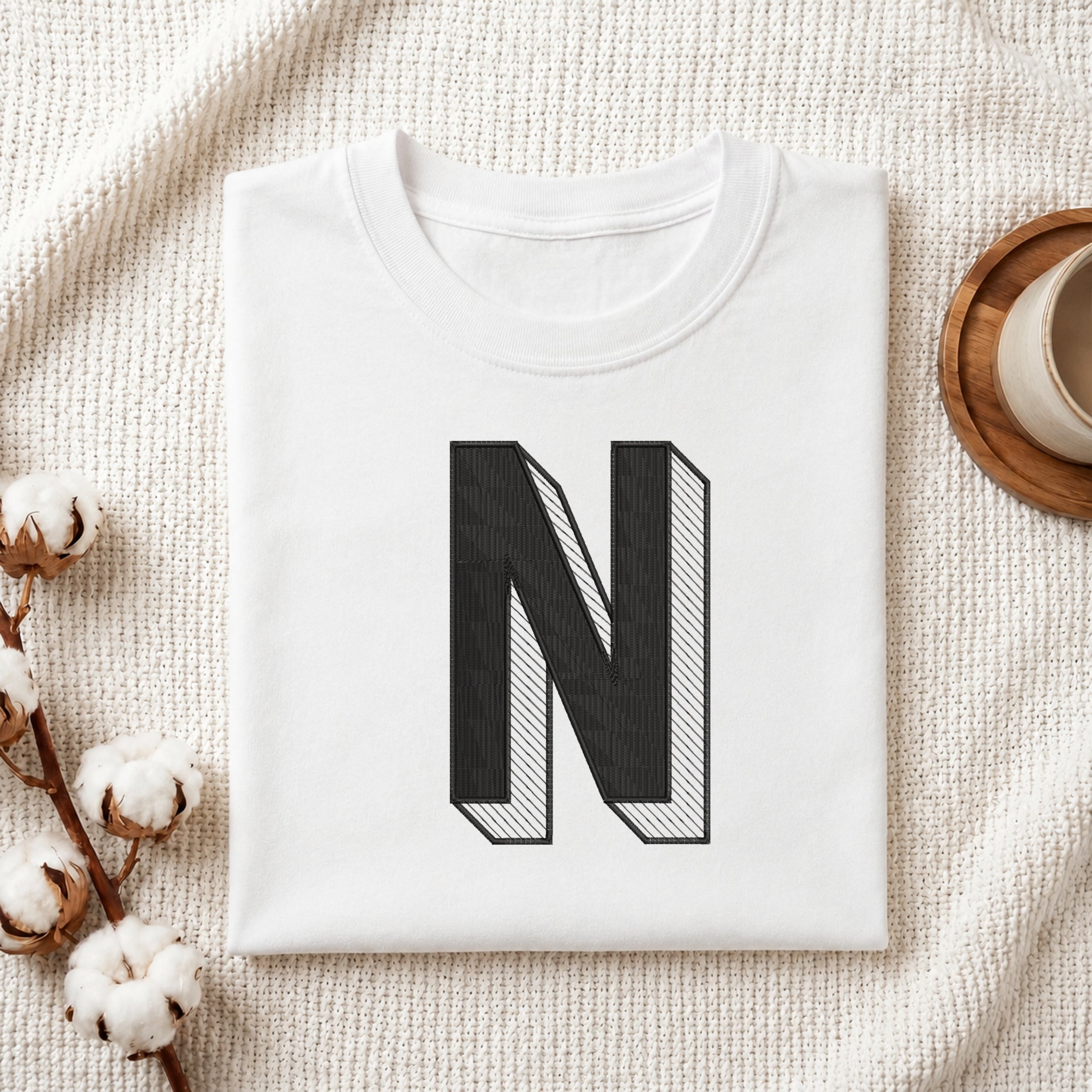 A folded white t-shirt with a large black block letter 'N' printed on the front, styled on a textured cream knit blanket with cotton stems and a cup in the background.