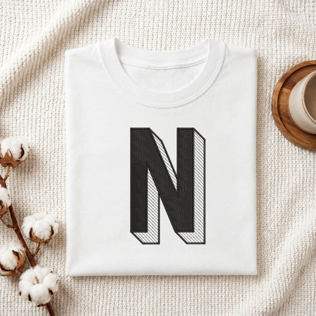 A folded white t-shirt with a large black block letter 'N' printed on the front, styled on a textured cream knit blanket with cotton stems and a cup in the background.