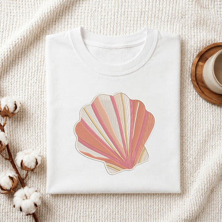Folded white cotton t-shirt mockup featuring a pink and beige coastal seashell machine embroidery design, styled with cotton stems and a wooden cup.