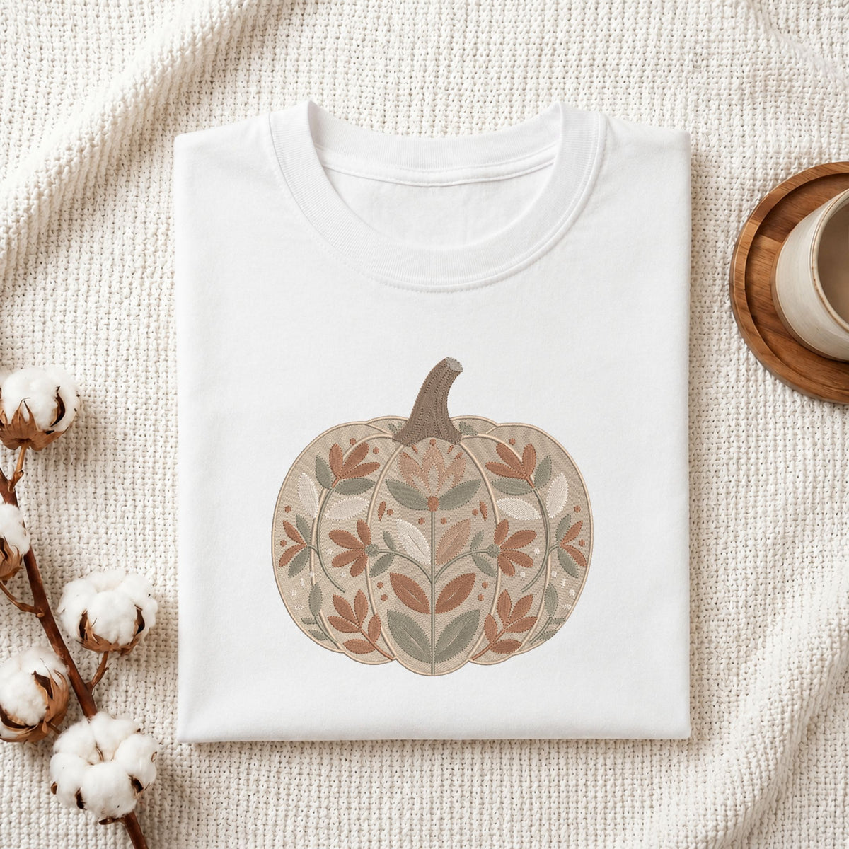 A folded white crewneck t-shirt with a stylized autumn pumpkin graphic, displayed on a textured cream fabric background with cotton bolls and a wooden cup on a coaster.