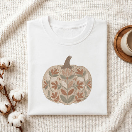 A folded white crewneck t-shirt with a stylized autumn pumpkin graphic, displayed on a textured cream fabric background with cotton bolls and a wooden cup on a coaster.