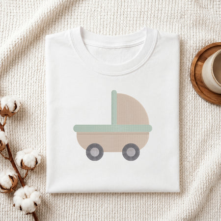 A folded white baby t-shirt with a minimalist pastel stroller graphic, styled on a textured cream knit blanket with cotton bolls and a wooden cup.