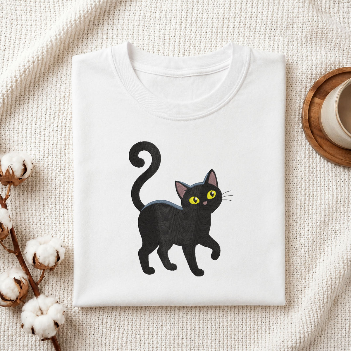 A folded white crewneck t-shirt featuring a cartoon illustration of a black cat with yellow eyes, styled on a textured cream fabric with cotton bolls and a wooden cup.