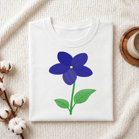 A folded white crewneck t-shirt featuring a graphic of a blue flower with green leaves and a yellow center, styled on a textured cream fabric with cotton bolls and a wooden cup.