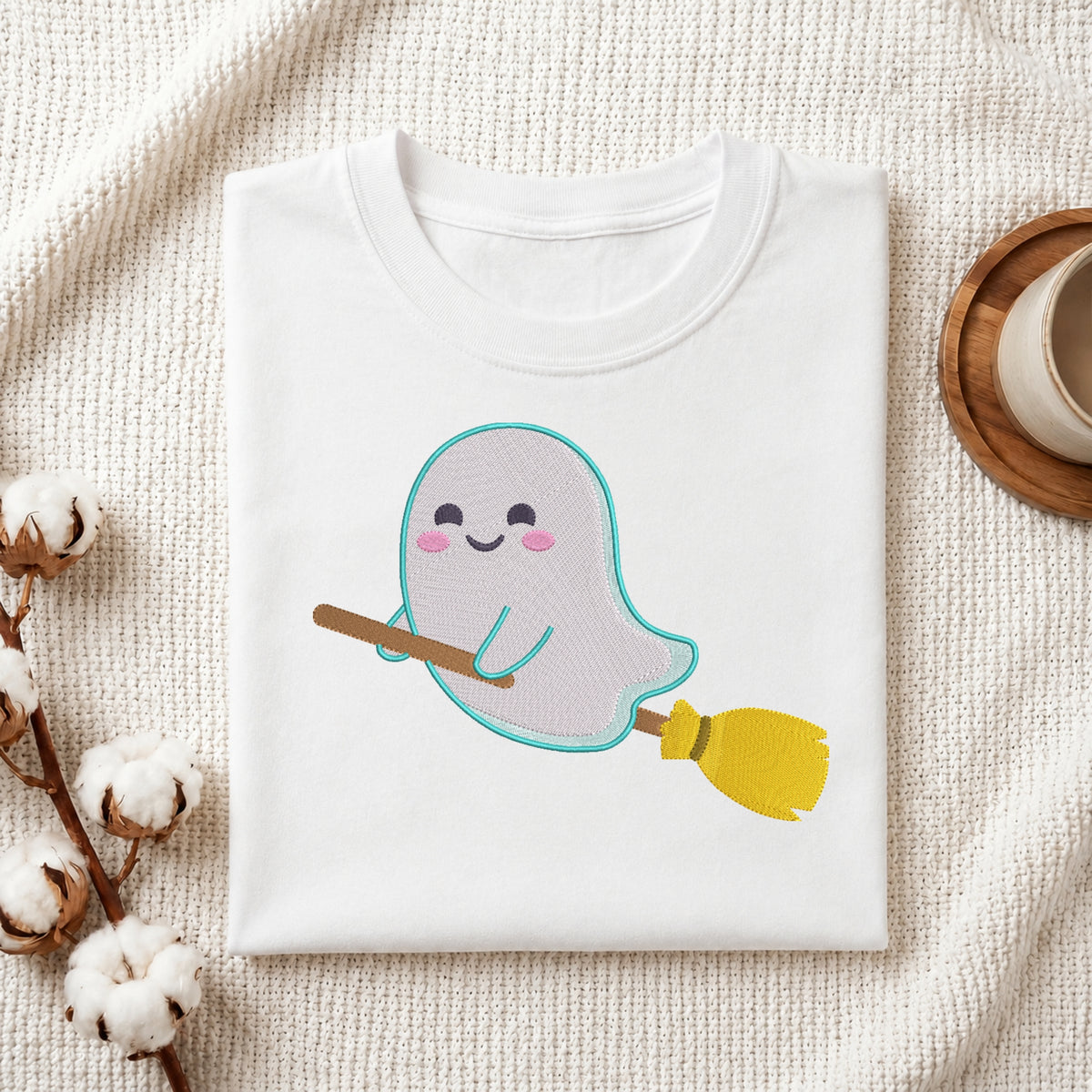 A folded white crewneck t-shirt featuring a cute cartoon ghost riding a yellow and brown broomstick, styled on a textured cream fabric background with cotton bolls and a coffee cup.