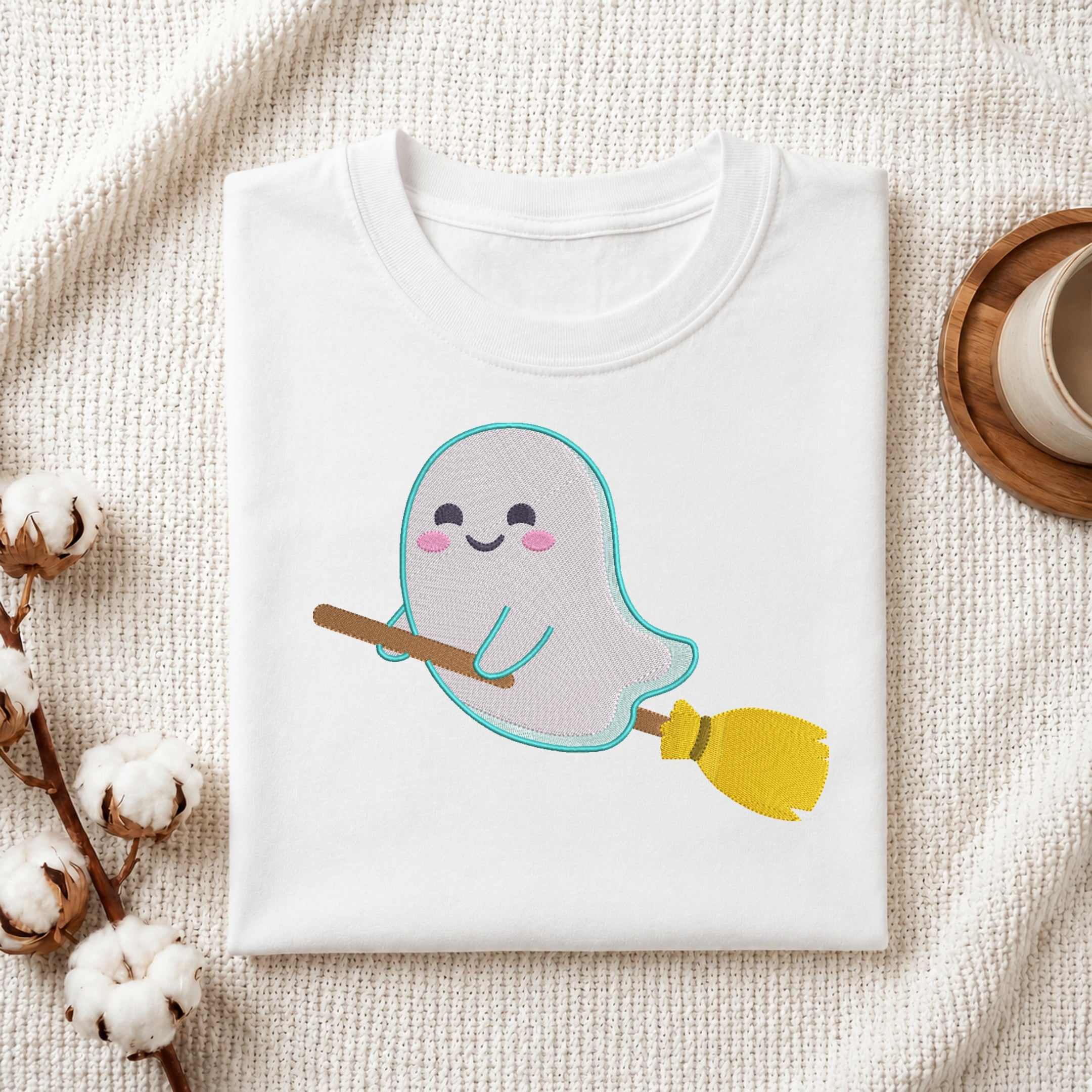 A folded white crewneck t-shirt featuring a cute cartoon ghost riding a yellow and brown broomstick, styled on a textured cream fabric background with cotton bolls and a coffee cup.