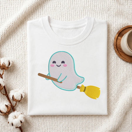 A folded white crewneck t-shirt featuring a cute cartoon ghost riding a yellow and brown broomstick, styled on a textured cream fabric background with cotton bolls and a coffee cup.