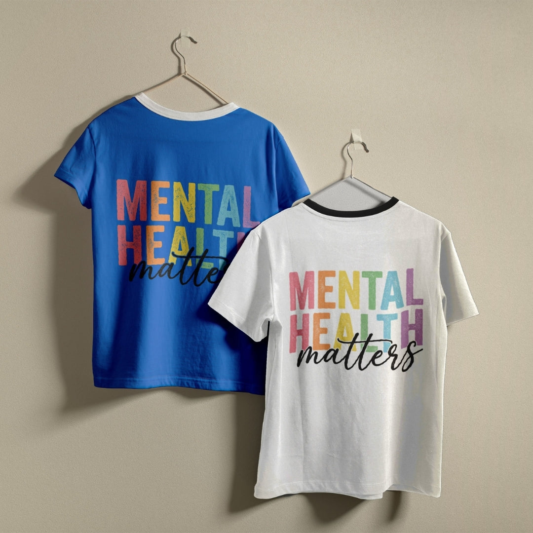 Two ringer t-shirts on hangers against a beige wall, showing the front and back designs. The blue shirt shows the back with 'MENTAL HEALTH matters' in rainbow block letters and black script.