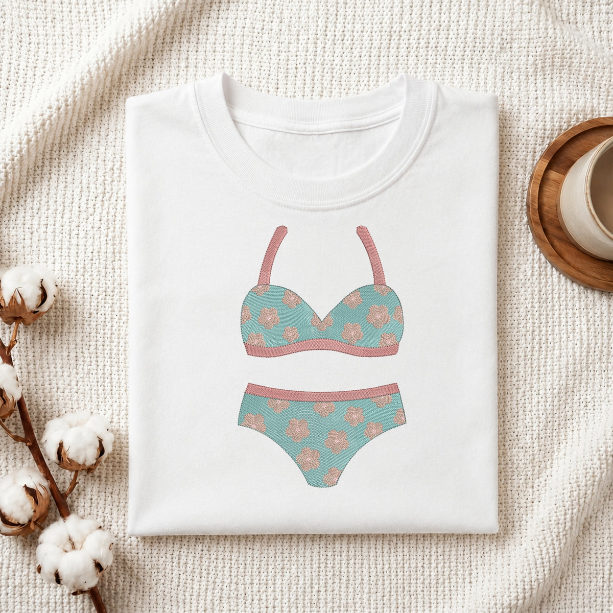 A folded white T-shirt with a printed bikini graphic in teal and dusty pink, styled on a textured cream fabric with cotton bolls and a small cup on a wooden tray.