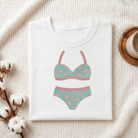 A folded white T-shirt with a printed bikini graphic in teal and dusty pink, styled on a textured cream fabric with cotton bolls and a small cup on a wooden tray.