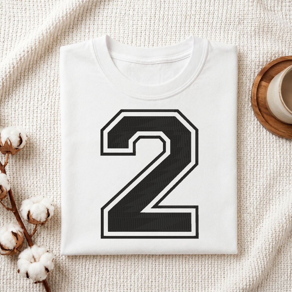 A folded white T-shirt with a large black number 2 outlined in white on the front, styled on a textured cream fabric with cotton stems and a small cup at the side.