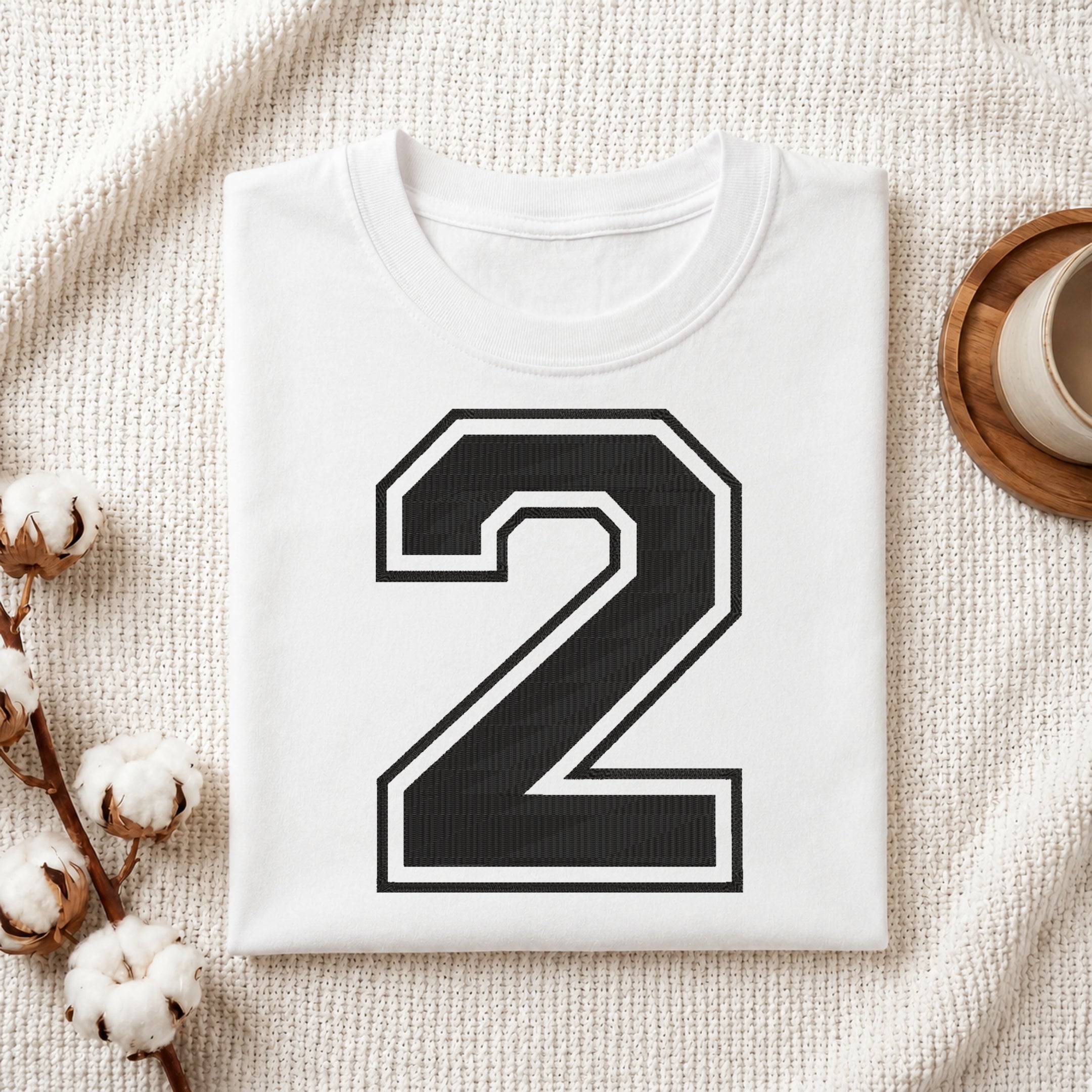 A folded white T-shirt with a large black number 2 outlined in white on the front, styled on a textured cream fabric with cotton stems and a small cup at the side.
