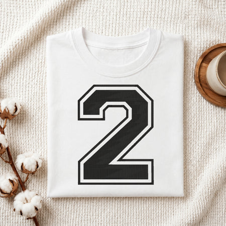A folded white T-shirt with a large black number 2 outlined in white on the front, styled on a textured cream fabric with cotton stems and a small cup at the side.