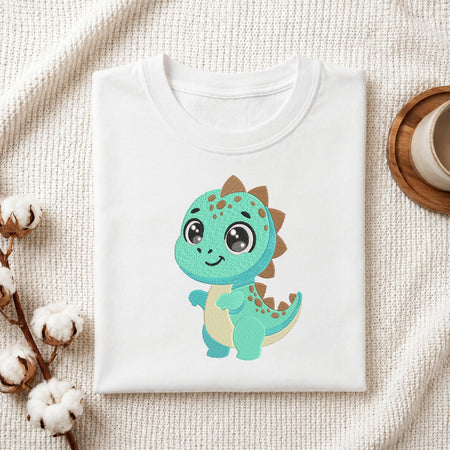 A folded white crewneck t-shirt featuring a cute, glitter-textured teal and cream dinosaur graphic on the front, styled on a textured cream fabric with cotton bolls and a wooden cup.