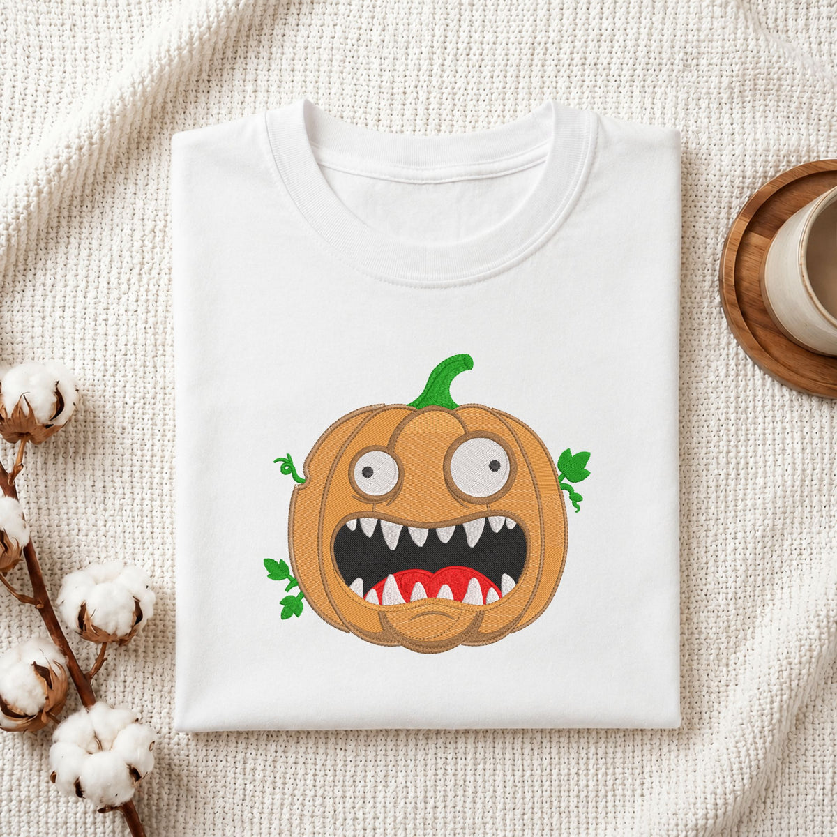 A folded white t-shirt with a cartoon monster pumpkin graphic, styled on a textured cream fabric with cotton bolls and a wooden cup.