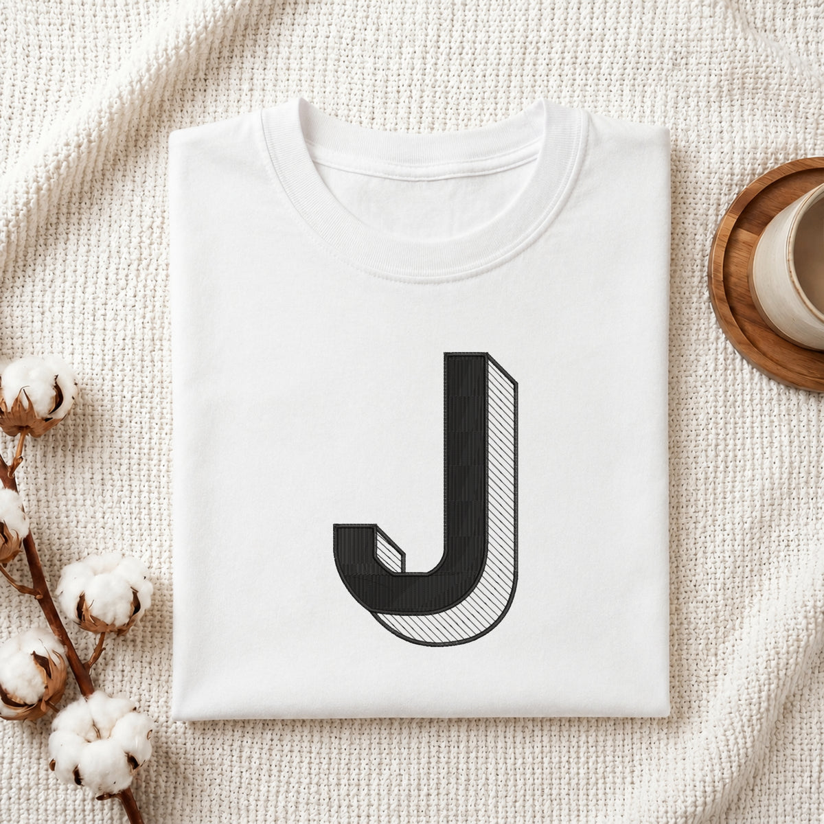 A folded white t-shirt with a large black graphic letter 'J' printed on the front, styled in a flat-lay on a textured cream fabric with cotton bolls and a wooden cup.