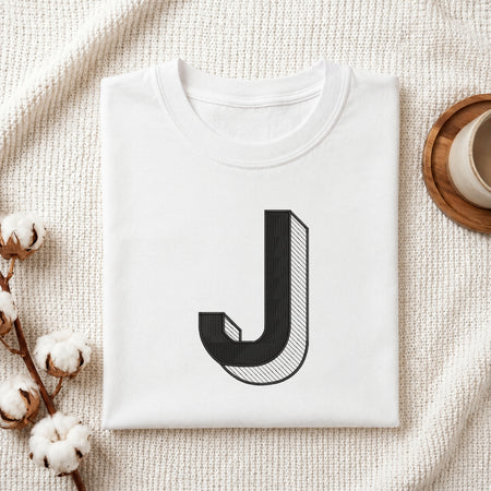 A folded white t-shirt with a large black graphic letter 'J' printed on the front, styled in a flat-lay on a textured cream fabric with cotton bolls and a wooden cup.