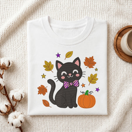 A folded white crewneck t-shirt with a cute black cat graphic wearing a purple striped bow tie, surrounded by autumn leaves, stars, and a pumpkin, styled on a textured cream fabric with cotton stems and a wooden cup.