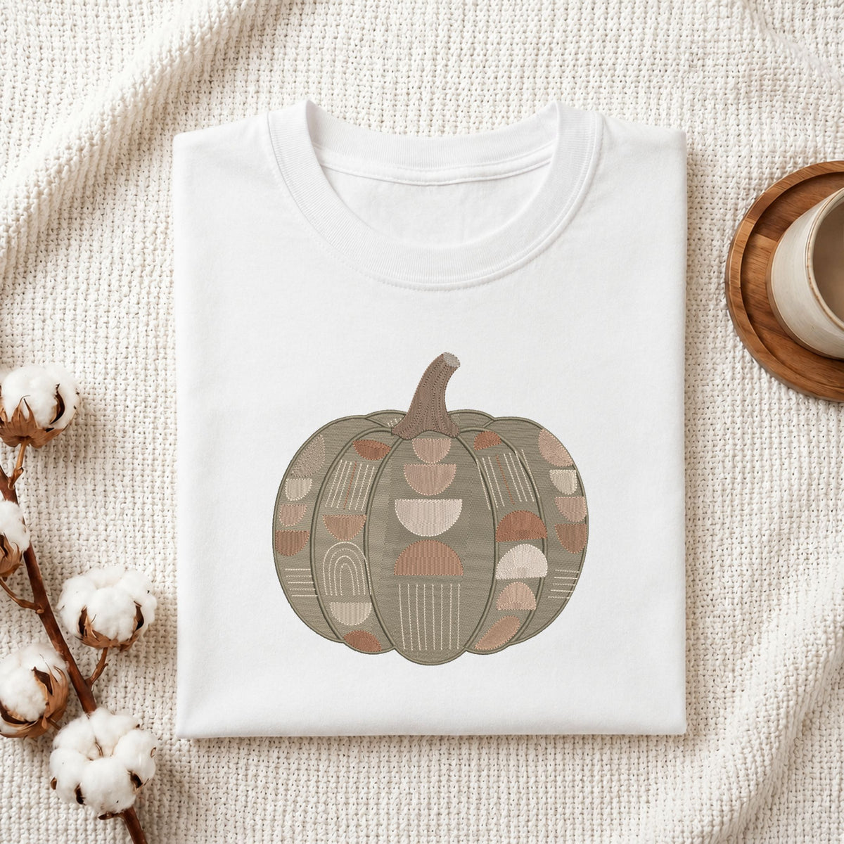 Folded white cotton t-shirt mockup featuring a geometric pumpkin machine embroidery design, styled with cotton stems and a wooden cup.