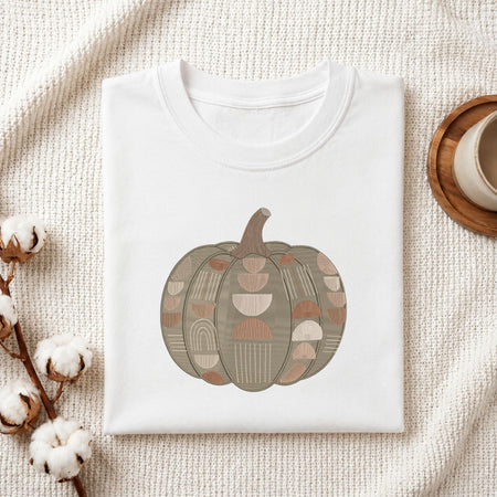 Folded white cotton t-shirt mockup featuring a geometric pumpkin machine embroidery design, styled with cotton stems and a wooden cup.