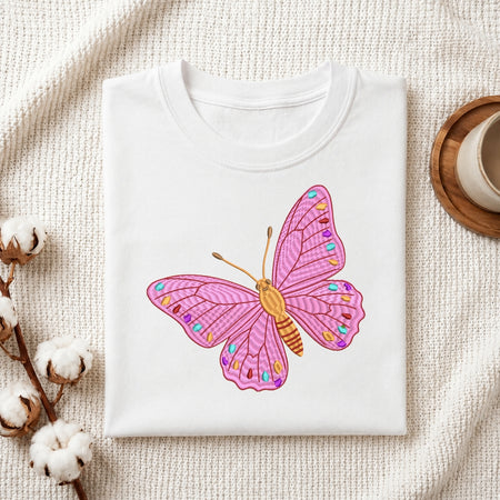 A white folded t-shirt featuring the embroidered pink butterfly design, displayed on a textured white fabric background with cotton sprigs and a wooden coaster.